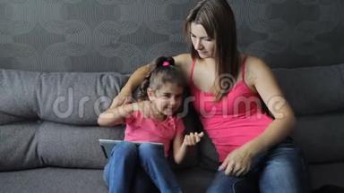 母亲带着孩子坐在沙发上用平板电脑。 女人会让坐在沙发上的婴儿痒痒。 在训练中吐口水。